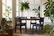 ambiente com cadeira para mesa de jantar uma preta ao redor de uma mesa retangular em tom de madeira e plantas verdes ao redor